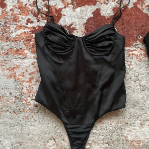 Svart korsettbody med tunna axelband - Snygg svart korsettbody med tunna axelband och markerade kupor. Stilren design med lätt rynkade detaljer vid bysten och figurnära passform. Perfekt att styla till både jeans och kjol .