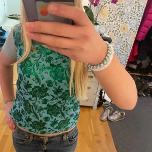 Grön blommig topp från Villain - Säljer en unik topp från Villain i storlek S. Toppen har grå korta ärmar och en glittrig grön blommig framsida i syntetmaterial. Perfekt för festliga sommarkvällar. Priset kan diskuteras.