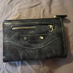 Svart plånbok med dragkedja och detaljer - Snygg svart plånbok i skinn med guldiga metalldetaljer och dragkedja framtill. Plånboken har flera kortfack och ett fack med tryckknapp. Perfekt storlek för att få plats med det viktigaste.