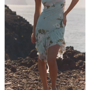 Blommig asymmetrisk klänning med volang - Superfin klänning med blommigt mönster i ljusa pastellfärger. Klänningen har en asymmetrisk nederkant med volangdetaljer och smala axelband. Perfekt för sommarens alla tillfällen. Materialet känns lätt och luftigt.