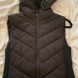 Svart dunväst från ZARA med huva - Säljer en svart dunväst från ZARA i storlek S. Västen har en hög krage, huva och dragkedja framtill. Perfekt att bära över en tröja för extra värme. Snyggt vadderad med diskret design.