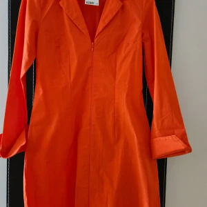 Orange långärmad klänning med dragkedja - Säljer en orange klänning med lång ärm och dragkedja framtill. Klänningen har krage och en rak passform, perfekt för dig som gillar färgstarka plagg.