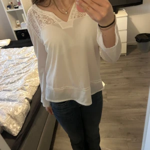 Vit blus med spetsdetaljer - Säljer en vit blus med långa ärmar och v-ringning. Blusen har fina spetsdetaljer vid axlar och över bröstet samt dekorativa sömmar nertill. Perfekt för en stilren och feminin look.