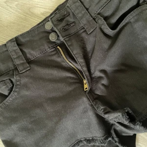 Svarta jeansshorts med fransig kant - Svarta jeansshorts med rå fransig kant nertill, dragkedja och dubbla knappar i midjan. Klassisk femficksmodell med bälteshällor och bakfickor. Perfekta för en avslappnad stil.🥰💗🤘