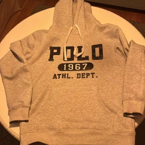Grå hoodie från Polo Ralph Lauren - Säljer en grå hoodie från Polo Ralph Lauren med svart tryck framtill: 'POLO 1967 ATHL. DEPT.'. Tröjan har huva med dragsko och en stor magficka. Perfekt för en avslappnad stil.