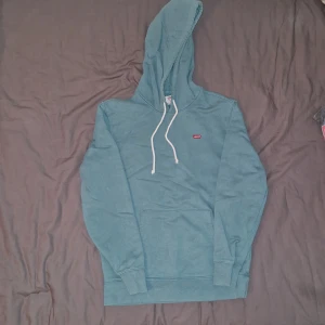 Turkos hoodie från Levi's - Säljer en ljusblå hoodie från Levi's med vit dragsko och klassisk logga på bröstet. Tröjan har känguruficka framtill och ribbade muddar vid ärmslut och nederkant. Perfekt för en avslappnad stil. Vet inte hur gammal den är eftersom den är min brorsas gamla, men den är ganska oanvänd och har inga täcken på användning heller 😊