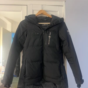 Svart vinterjacka från Sali Racing - Säljer en svart pufferjacka från Sea Racing med huva och dragkedja framtill. Jackan har en sportig look med logga på bröstet och ärmen. Perfekt för kyliga dagar och har en längre passform baktill.