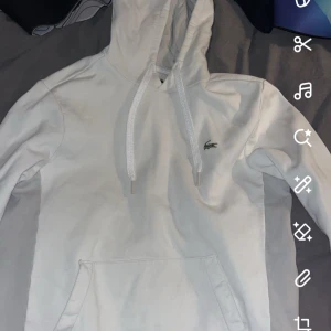 Vit hoodie från Lacoste - Säljer en stilren vit hoodie från Lacoste med klassisk krokodillogga på bröstet. Tröjan har huva med dragsko och en stor magficka framtill. Perfekt för en avslappnad look.
