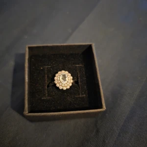 Ring med Aquamarin  - Elegant ring med en oval klar sten i mitten, omgiven av små gnistrande diamanteri en halo-design. Ringen har en klassisk och tidlös look som passar perfekt till speciella tillfällen. 