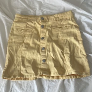 Gul kortkjol med knappar från Lindex - Säljer en ljusgul kort jeanskjol från Lindex med silverfärgade knappar framtill och två dekorativa fickor. Kjolen har en rak passform.
