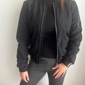 Svart bomberjacka - Säljer en svart bomberjacka med dragkedja framtill och ribbade muddar vid ärmslut och midja. Jackan har lång ärm och klassisk bombermodell, perfekt för en cool och avslappnad stil.