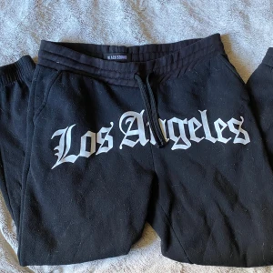 Svarta mjukisbyxor med Los Angeles-tryck från  NewYorker i modellen BLACK SQUAD  - Svarta mjukisbyxor från Black Squad med stort vitt Los Angeles-tryck framtill. Byxorna har resår och snörning i midjan samt en bakficka. Perfekta för en avslappnad stil. 