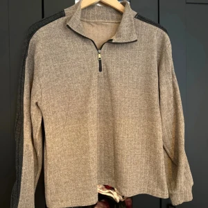 Beige tröja med dragkedja och ståkrage - Säljer en beige långärmad tröja med halv dragkedja och ståkrage. Tröjan har mörkgrå detaljer längs axlar och ärmar, vilket ger en sportig look. Perfekt för kyliga dagar och enkel att matcha med olika outfits.