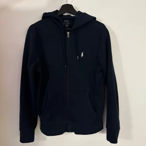 Mörkblå hoodie från Polo Ralph Lauren - Säljer en mörkblå hoodie från Polo Ralph Lauren med dragkedja och huva. Tröjan har en liten vit broderad logga på bröstet och två fickor framtill. Perfekt för en avslappnad stil.