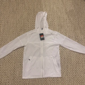 nike windbreaker - Säljer en stilren vit vindjacka från Peak Performance med huva och dragkedja framtill. Jackan har långa ärmar och två fickor med dragkedja. Perfekt för blåsiga dagar och enkel att matcha med olika outfits. Står M men sitter som S