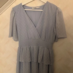 Ljusblå prickig klänning från NA-KD - Supersöt ljusblå klänning från NA-KD med vita prickar, v-ringning och smock i midjan. Klänningen har korta ärmar och volangdetaljer vid kjolen. Perfekt för sommarens alla tillfällen! (Har aldrig används)