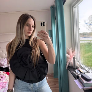 Svart ärmlös topp med stor volangdetalj Magda Butrym H&M M storlek  - Unik svart ärmlös topp med en stor, rund volangdetalj framtill. Toppen har en hög halsringning och är tillverkad i ett slätt material. Perfekt för dig som vill sticka ut med en cool och trendig look.