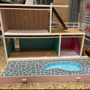 Dockskåp med pool och kök - Säljer ett stort dockskåp i trä med två våningar, färgglada tapeter, trappa och altan. Uteplats med pool och stengolvsmönster. Medföljer ett detaljerat kök i miniatyr med skåp, ugn och blåvit kakelvägg. Perfekt för docklekar och inredningsfantaster.