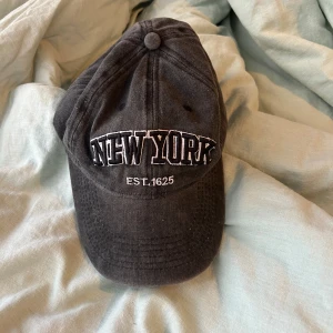 Svart keps med New York-brodyr - Svart keps med böjd skärm och broderad text 'NEW YORK EST. 1625' framtill. Justerbar rem baktill i samma tyg. Klassisk och stilren design, perfekt för en avslappnad look.