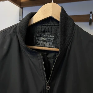 Svart bomberjacka från Levi's - Snygg svart bomberjacka från Levi's med klassisk ribbad krage och dragkedja framtill. Jackan har två fickor med knappstängning och en stilren design som passar till många outfits.