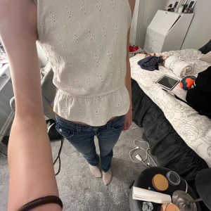 Vit ärmlös blus med broderade detaljer - Säljer en söt vit blus med broderade hålmönster och volang nertill. Blusen är ärmlös och har en lätt utsvängd passform. Perfekt till jeans eller kjol för en fräsch look. Skönt tyg som gör den väldigt bekväm. Storleken är 146/152 på barnavdelningen men passar som xs/xss