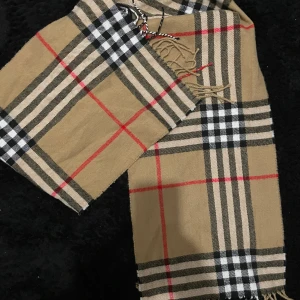 Beige rutig halsduk - Snygg beige halsduk med klassiskt rutigt mönster i svart, vitt och rött. Halsduken har fransar i ändarna och passar perfekt för att ge din outfit en stilren touch.