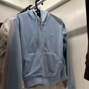 Ljusblå velour hoodie från Juicy Couture - Säljer en ljusblå hoodie i mjuk velour från Juicy Couture. Tröjan har dragkedja, huva med snören och två fickor framtill. Ikonisk JC-detalj på dragkedjan och broderad logga på bröstet. Perfekt för en avslappnad och trendig look.