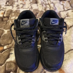 Svarta Nike Air Max sneakers - Säljer ett par svarta Nike Air Max sneakers med reflekterande silverdetaljer och klassisk Nike-logga på sidan och hälen. Skorna har snörning och en synlig Air Max-dämpning i sulan för extra komfort. Perfekta för en sportig och stilren look.