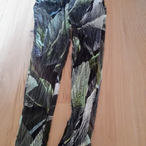 Nwt Mönstrade byxor från Lindex storlek S - Säljer ett par tunna, mönstrade byxor från Lindex i gröna, svarta och vita nyanser. Byxorna har ett unikt abstrakt mönster och är tillverkade i viskos med lite elastan för stretch. Djungelmönster. Djungel blad, löv. Perfekta för dig som gillar statement-plagg!