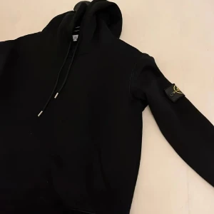 Svart hoodie från Stone Island - Säljer en svart hoodie från Stone Island med klassisk patch på ärmen. Tröjan har huva med dragsko och ribbade muddar. Perfekt för dig som gillar streetwear och vill ha något stilrent men ändå med attityd.