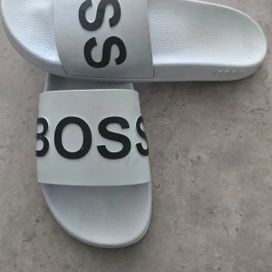 Vita sandaler från BOSS - Säljer ett par vita sandaler från BOSS med bred rem och stor svart logga över foten. Perfekta för sommaren och enkla att ta på sig. Klassisk design med platt sula och öppen tå.