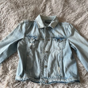 Ljusblå jeansjacka med slitningar - Säljer en klassisk ljusblå jeansjacka med slitna detaljer på bröstfickan och framtill. Jackan har knappar framtill, två bröstfickor med lock och lång ärm. Perfekt för våren och sommarn 