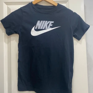 Svart t-shirt från Nike med logga - Säljer en svart t-shirt från Nike med stor vit logga och text på bröstet. Klassisk rund halsringning och korta ärmar. Perfekt för en sportig och avslappnad stil. I storlek M. 