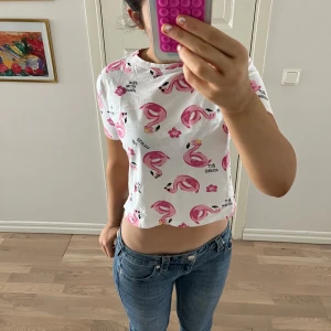 Vit t-shirt med flamingomönster - Söt vit t-shirt med rosa flamingos och blommor. Perfekt för sommaren med text som 'Summer Vacay Mood'. Kortärmad och i mjukt material.