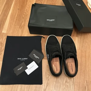 Svarta sneakers från Saint Laurent - Svarta sneakers från Saint Laurent med vit logga på sidan och vit platt sula. Klassisk design med snörning och rund tå. Skorna har en stilren look och har detaljer i svart och vitt. Storlek 44-45. Använda några gånger pris kan diskuteras. Slitningarna ser bra ut. 