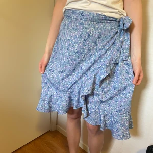 Blommig omlottkjol med volang🩵 - Säljer en blå och lila blommig omlottkjol med volangkant och knyt i midjan. Kjolen har en luftig känsla och är perfekt för varmare dagar. Mönstret är småblommigt och tyget känns mjukt mot huden. Perfekt till kanske midsommar eller en skolavslutning🌸