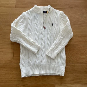 Quarter zip från Ralph Lauren - Vit kabelstickad tröja från Polo Ralph Lauren med halv dragkedja och broderad logga på bröstet. Tröjan har lång ärm, ribbade muddar och hög krage. Perfekt för lager-på-lager och klassisk stil.