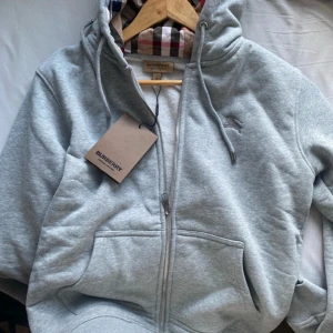 Burberry full zip - Säljer en stilren ljusgrå hoodie från Burberry med dragkedja och klassiskt rutigt foder i huvan. Tröjan har fickor framtill och en broderad logga på bröstet. Perfekt för en avslappnad och trendig look. Passar S också.Pris kan diskuteras vid snabb affär 