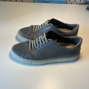 Grå sneakers från Axel Arigato - Säljer ett par stilrena grå sneakers från Axel Arigato med vit platt sula och klassisk snörning. Skorna har en rund tå och är tillverkade i skinn. Box och påse följer med.