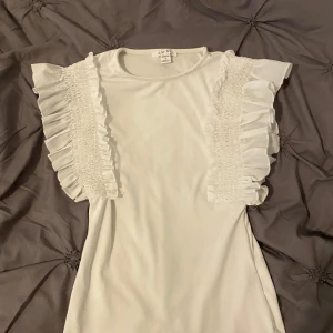 Vit topp med volangdetaljer - Säljer en vit topp med korta ärmar och breda volangdetaljer längs axlarna. Toppen har en rund halsringning och är tillverkad i ett mjukt material. Perfekt för dig som gillar feminina detaljer och enkel stil.