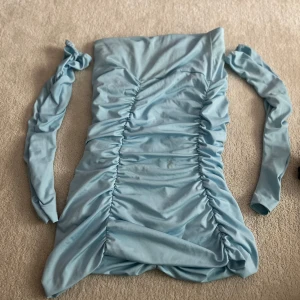 Ljusblå offshoulder klänning med rynkade detaljer - Trendig ljusblå klänning med offshoulder-design och långa ärmar. Klänningen har rynkade detaljer längs hela framsidan och är figurnära med kort längd. Perfekt för dig som vill sticka ut med en cool och modern look.