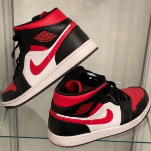 Nike Air Jordan 1 Mid röd, svart och vit - Säljer ett par Nike Air Jordan 1 Mid med klassisk färgkombination i rött, svart och vitt. Skorna har svart snörning, vit sula och röd Nike swoosh. Perfekta för dig som gillar streetwear och sneakers med ikonisk design.