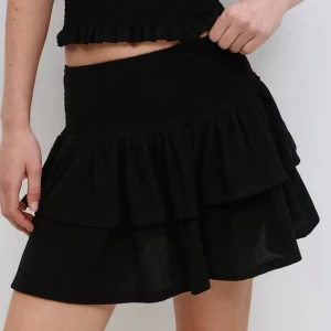 Svart volangkjol med resårmidja - En stilren och bekväm kjol som jag endast använt en gång. Kjolen har en fin passform och passar både till vardags och fest. Inga fläckar eller slitningar – ser ut som ny. (FRAKT INGÅR EJ)