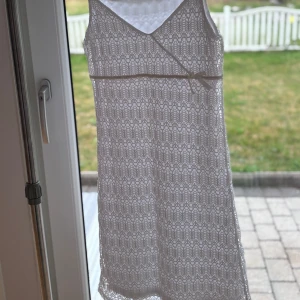 Vit spetsklänning med rosett - Vit klänning i spets med smala axelband och v-ringning. Klänningen har ett fint spetsmönster över hela och en liten söt rosett i midjan. 