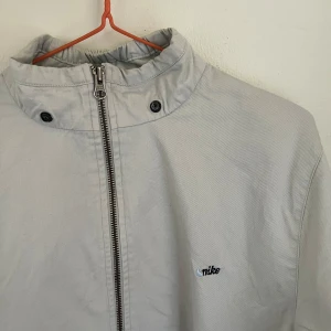 Ljusblå bommulsjacka från Nike - Nike bommulsjacka med minimalistisk design och diskret logga på bröstet. Jackan har hög krage, hel dragkedja framtill, två stora fickor och knappar vid ärmslut. Lite fläckig vid armarna(se bilder). 
