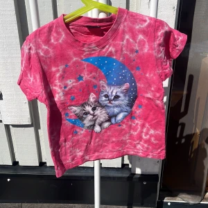 Rosa tie-dye t-shirt med kattmotiv VXO - Söt rosa tie-dye t-shirt från VXO med korta ärmar och ett stort tryck av två katter på en blå måne med stjärnor. T-shirten har en klassisk passform och är gjord i mjuk bomull, perfekt för dig som gillar gulliga och unika plagg.