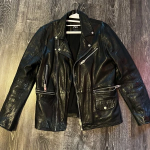 Svart skinnjacka från Zara - Snygg svart skinnjacka från Zara med klassisk bikerstil. Jackan har flera silvriga dragkedjor, snedställd front och coola detaljer som tryckknappar vid kragen. Perfekt för dig som vill ha en edgy look.