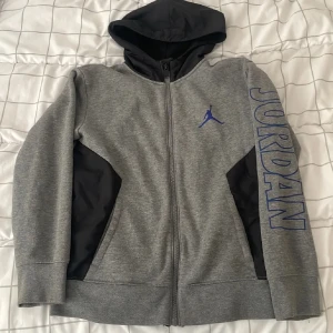 Grå Jordan hoodiejacka med blå logga - Snygg grå hoodiejacka från Jordan med svart huva och svarta detaljer vid sidorna. Stor blå Jumpman-logga på bröstet och JORDAN tryckt längs ena ärmen. Jackan har dragkedja hela vägen och är perfekt för dig som gillar sportig stil.