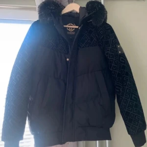 Svart pufferjacka Glorious Gangsta - Snygg svart pufferjacka från Glorious Gangsta med mönstrade detaljer och stor fuskpäls på huvan. Jackan har dragkedja, broderad logga och ett unikt geometriskt mönster på axlar och ärmar. Perfekt för kalla dagar när du vill sticka ut.