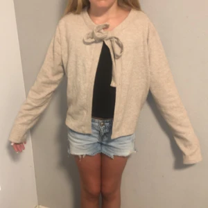 Beige kofta med knyt från 157, XS - Superfin beige kofta från 157 i mjukt material. Den har långa ärmar och en söt knytning vid halsen som ger en chill och trendig look. Perfekt att slänga över en topp eller klänning när det är lite kyligt ute.